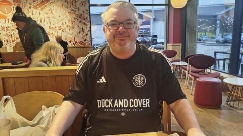 Mark Sharpley has greying hair, dark spectacles and is wearing the dark change kit of Macclesfield FC withshirt sponsor Duck and Cover emblazoned on it. He is sitting in a branch of Costa Coffee.