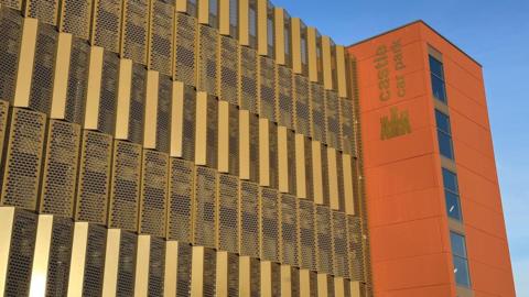 A multi-storey car park, with gold panelling and a terracota-coloured cladded stairwell to the right. There is a gold image of a castle underneath lettering which reads Castle Car Park.