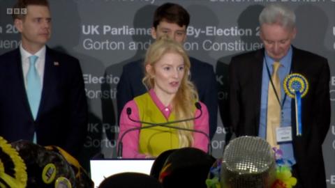 A blonde woman in a pink shirt and lime green sweater vest is seen behind a podium.