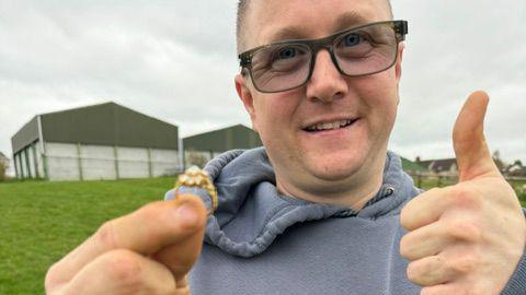 A man in his early 40s smiles as he stands in a field on a cloudy day. In one hand he is holding up a gold diamond ring, while he gives a thumbs up with the other hand. He is wearing a grey hoodie and black-rimmed glasses, the lens of which are slightly tinted. There are large metal barns behind him.