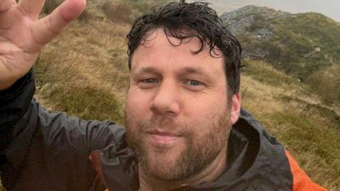 Selfie style picture of a bearded man with dark wavy hair on a hill. He is wearing a black and orange jacket and waving at the camera