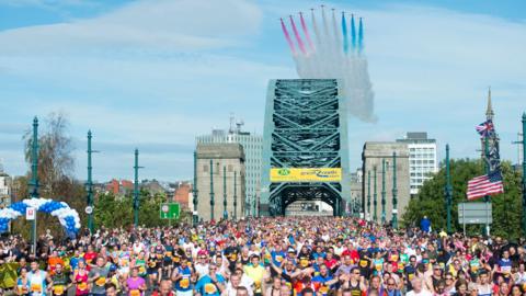 Crowds of runners, wearing colourful tops, run across the Tyne Bridge. A set of planes (Red Arrows) can been in the blue sky, leaving a trail of white, red and blue markings in the sky.