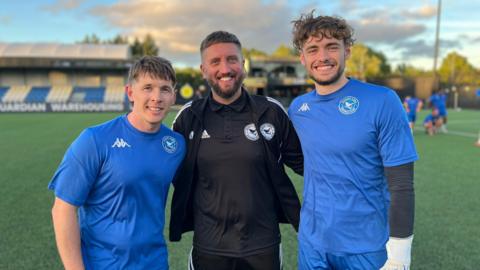 Three men in football kits smile at the camera. On the left and the right the men wear blue, and the man in the middle wears a black kit. In the background there is a grassy football pitch and football stands.