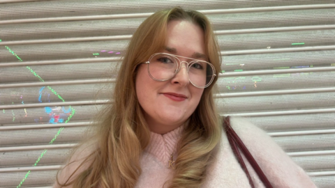 A woman with long blond hair and glasses in a pale jumper in front of a white slatted wall