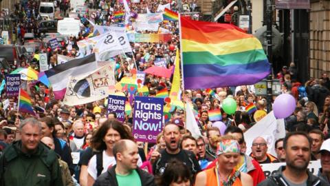 People in the Edinburgh Pride march walk along a road holding rainbow flags and trans rights signs 
