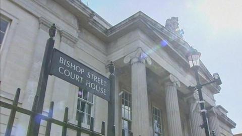 Bishop Street Courthouse, seen through iron railings. A sign with the buildings name is seen on the left while he entrance to the buildings four pillars are to the right.
