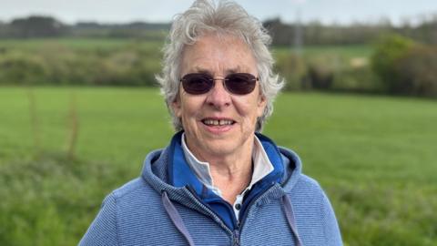 Kate has curly grey hair and round sunglasses. She wears a blue hooded top and stands in front of the fields where the solar farm will go. 