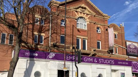 A three storey red brick building with white framed windows , a purple sign with white lettering saying Empire Gym & Studios, there is a tree in front of the building with no leaves on it.