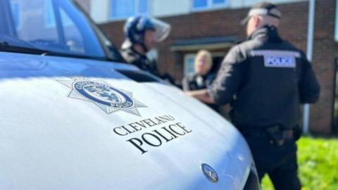 A silver police van in the foreground with the Cleveland Police logo on the bonnet. Three officers wearing dark clothing are talking to each other in the background.
