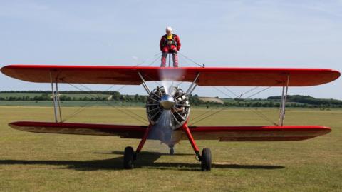 Harry Heasman, in a red and black jump suit, is shown atop a red biplane. The plane's singular propeller engine is shown in motion on a grass landing strip at the airfield.
