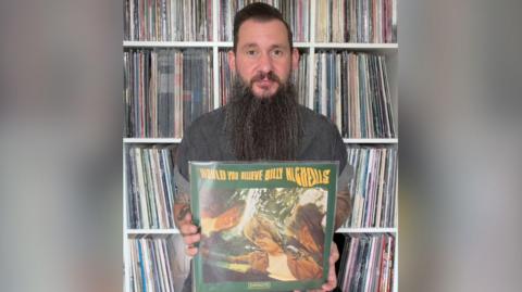 Martin is holding Billy Nicholls record, would you believe. The cover is green with yellow text. The image is a wavy effect photo of the main artists involved. Martin has dark hair and a very long beard. He is sat infant of loads of vinyl records in shelves 