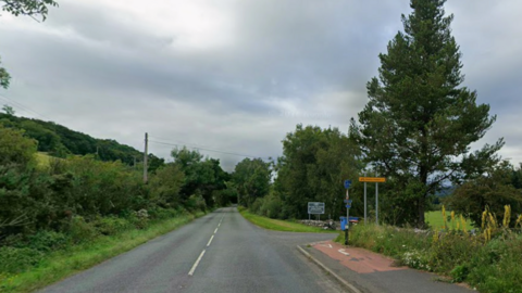 A straight road. There are green trees and hedges on either side. There is a road off to the right, with a few signs which are too far away to read. 