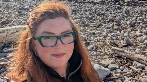 Close up of selfie of Sian smiling whilst stood on a pebbled beach. She has straight ginger hair and wears a black fleece and blue glasses. 