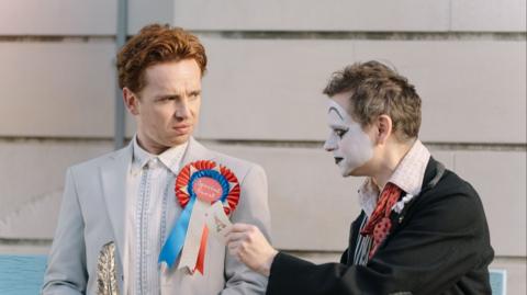 A clown pins a rosette on the jacket of a man.