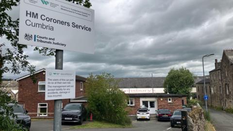 The coroner's office. It's a small single-storey brick building with cars parked outside of it. There's a sign saying HM Coroners Service Cumbria on a post.