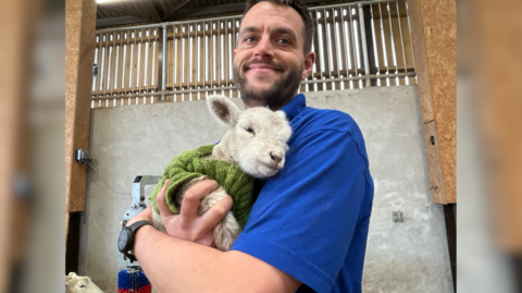 The image shows a man in a blue shirt holding a lamb in a small green jumper. They are in a barn and some fencing as well as the top of another lamb's head can be seen in the background.