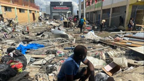 A man squats in the foreground as a swath of destruction sets out behind him ending at a destroyed grocery store.