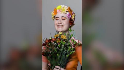 A young woman smiling, wearing a crown of flowers, orange eyeshadow and holding a bunch of carnations.