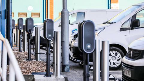 A fleet of white electric council vans hooked up to EV chargers.