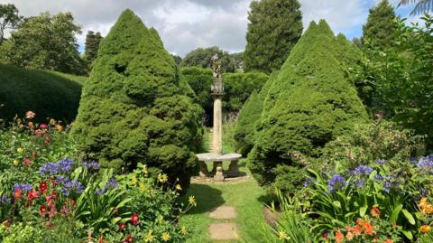 A walled garden view of a stone path between beds of colourful flowers leading to a stone bench with a tall plinth rising from it's centre. A wood pigeon sits on it's top. Rows of conical shaped spruce trees stand either side of the bench