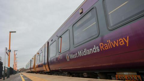 The side of train carriages pulled up on a platform. The words West Midlands Railway can be seen on one carriage