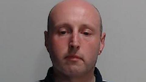 Ross Jameson mugshot. A bald man wearing a blue, white and yellow striped polo-shirt. 