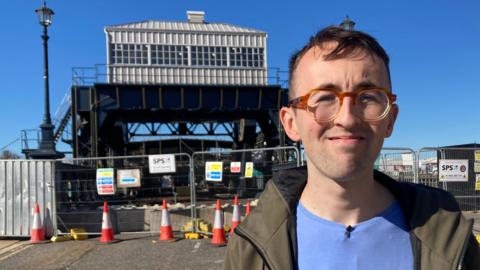 Laurence Gooch, a 28-year-old man with glasses and short brown hair, is wearing a light blue T-shirt and khaki jacket. He stands on a road in front of a 100-year-old bridge, which has dark steelwork and is topped by a control cabin made of white wood and small glass panels. The road and bridge are blocked with a metal fence and red and white cones.