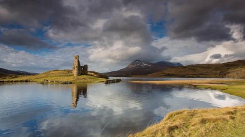 A view across a calm Loch Assynt towards the ruins of Ardvreck Castle. The ruins include a tower.