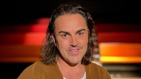 Stephen Crocker is in the middle of the frame. He has shoulder-length brown curly hair and is wearing a white T-shirt and a brown jacket. Behind him are rows of seats.