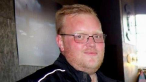 A man with short blond hair and stubble is pictured looking towards the camera while wearing a black motorcyclist jacket.