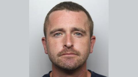 A police mugshot of Simon Orr. He is looking at the camera and has cropped brown hair, and a short moustache and beard.