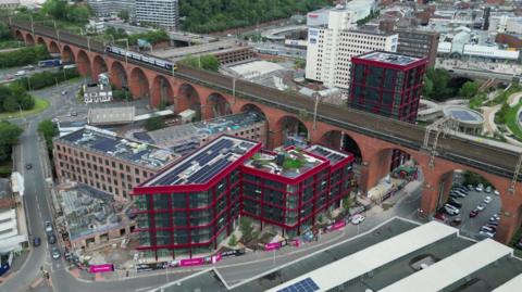 A drone image of Stockport town centre. There is a train travelling along the historic viaduct which spans the length of the picture. In the middle of a ring road is a number of new, modern high rises located next to former mill buildings.