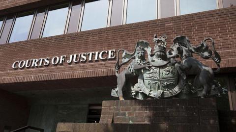 Portsmouth Crown Court. A red brick building with a row of windows above a metal sign reading "Courts of Justice". A large metal statue of an insignia sits to the right of the sign. 