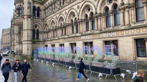 A multi-coloured sign (purple, green and orange) made out of metal and spelling out Bradford 2025 with the Yorkshire stone walls of Bradford City Hall behind it and people walking in front. 