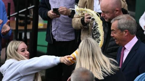 A woman in a grey hoody is holding a paper cup in her outstretched hand. There is yellow milkshake coming out of it, flying through the air in the direction of Nigel Farage. He is wearing a dark suit with pink shirt and tie.