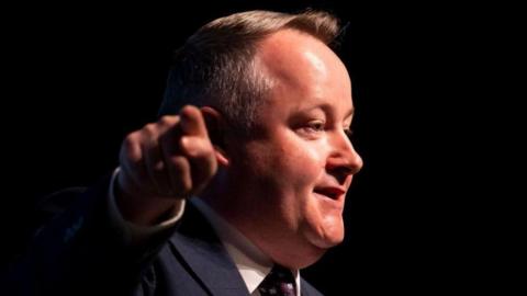 Darren Millar is pointing towards the camera while looking away at a right angle towards an audience. He is wearing a suit, tie and shirt, and is stood before a dark background.