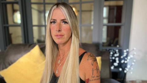 A woman with long blonde hair, wearing a black vested top with tattoos visible on one arm. 