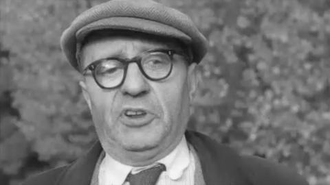 Black and white image of older man in flat cap and glasses grimacing