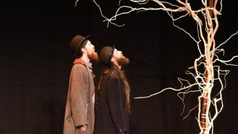 Two men look at at a tree which is white in colour and has no leaves. They stand in front of a black background. They both have beads, they wear bowler hats and long winter coats. 