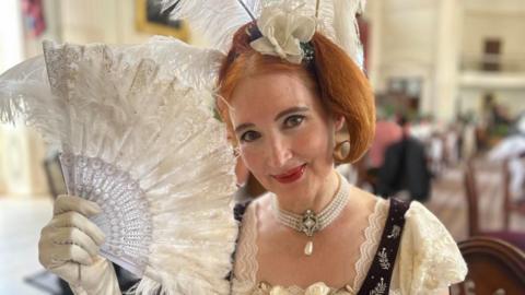 Jane Austen tour guide Theresa Roche is dressed in cream and purple Georgian clothing with gloves, a pearl necklace and a feathery head piece. She is holding an open fan in her right hand and she is smiling.