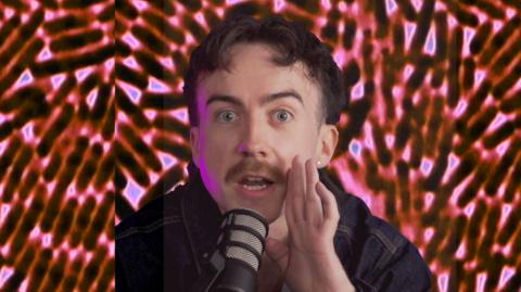 A man is holding his hand to his mouth and is speaking into a mic. He has dark brown short hair, blue eyes and a light brown moustache. He is wearing a dark denim jacket.The background is a close-up image of germs.