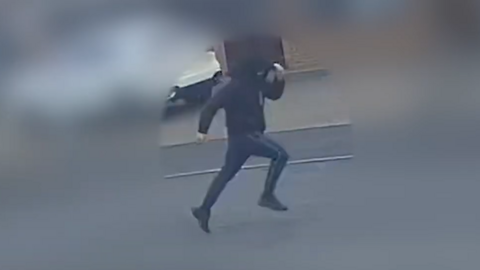 CCTV footage of a man wearing dark clothing. He appears to be running with his arms and legs stretched out. 