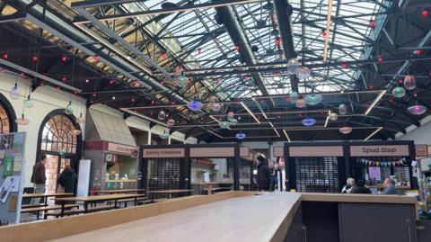 A photo of the Christmas lights at Lincoln's Cornhill Market. The coloured lightbulbs have been strung between metal rafters. Empty shops are visible in the background.