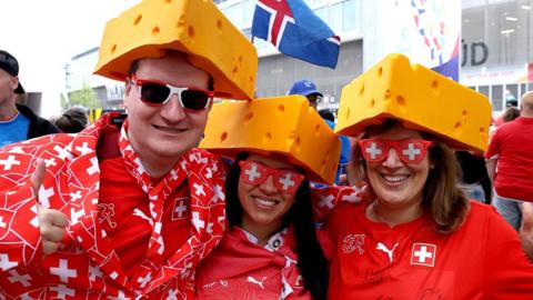Three Switzerland fans at Euro 2025 standing together wearing read and white outfits and glasses, and yellow triangular cheese hats