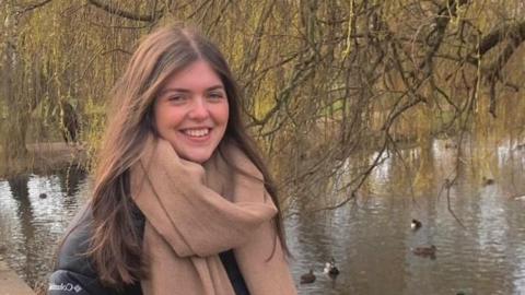 Nel Jones wearing a heavy coat and scarf, smiling in front of a pond with ducks 