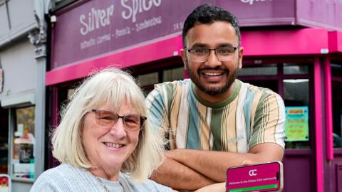 Councillor Maureen Cummings holding a phone showing the Citizen Coin app, pictured with Abrar Nawaz outside his Silver Spice sandwich and curry shop in Wakefield city centre