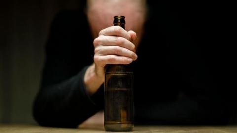 A man sitting alone at home at a table holding a bottle of beer in his hand. He is facing down.