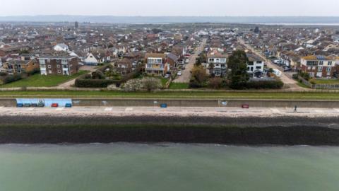 An aerial photo showing the sea in the foreground and rows of houses in the background. It is cloudy.