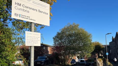 A general view of the access to the Coroners Court in Cockermouth. A sign reading "HM Coroners Service Cumbria" is in the foreground. Behind it is a small car park with a few cars parked in front of the court, which is a small red-brick building with a white PVC door.