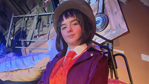 Young girl acting in a play wearing a bowl hat, purple blazer, orange waistcoat and tie with white shirt,  set in background 